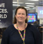 A woman with long brown hair, wearing a dark blue top and a lanyard, smiles at the camera in an office setting with digital displays in the background.