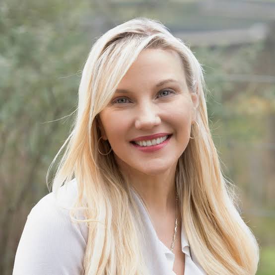 A woman with long blonde hair and a friendly smile, wearing a light-coloured top, standing outdoors with a blurred natural background.